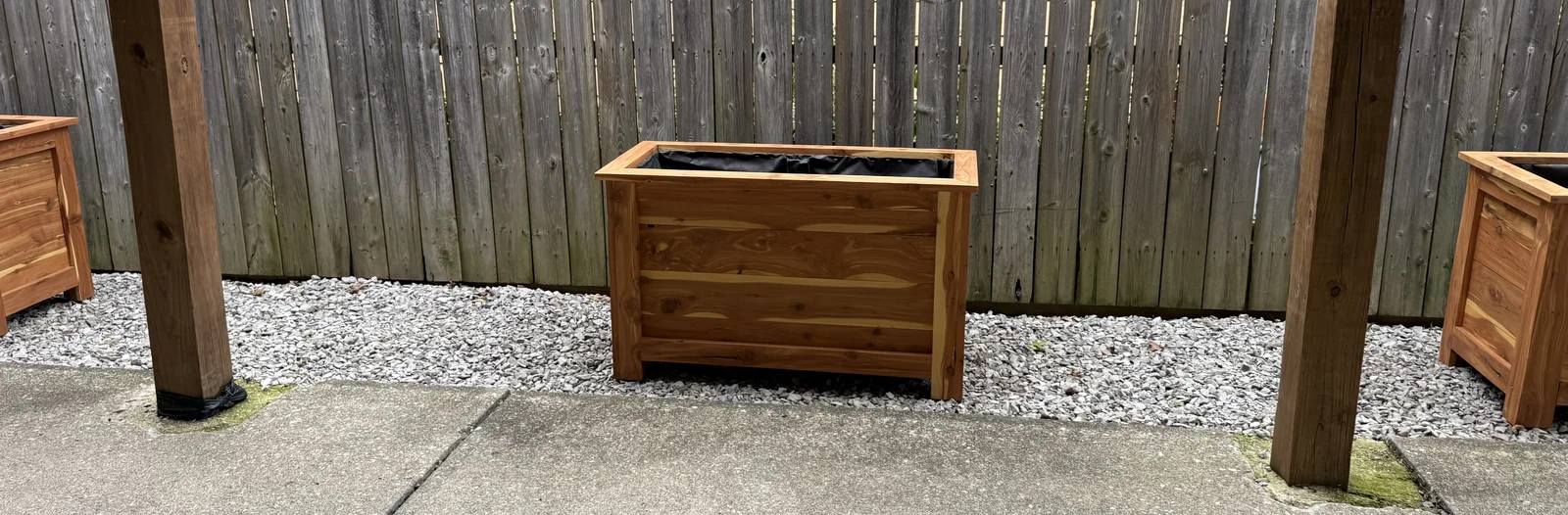 A delivered cedar planter centered between patio posts, with its framed sides and black liner visible.