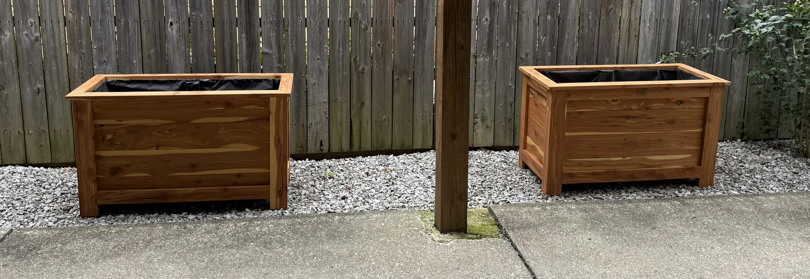 Two delivered cedar planters with black liners on gravel beside the fence.